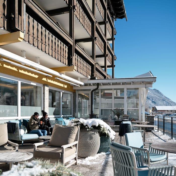 Hotelterrasse mit Wintermöbeln, zwei Personen trinken Kaffee bei Sonnenschein, Alpen im Hintergrund