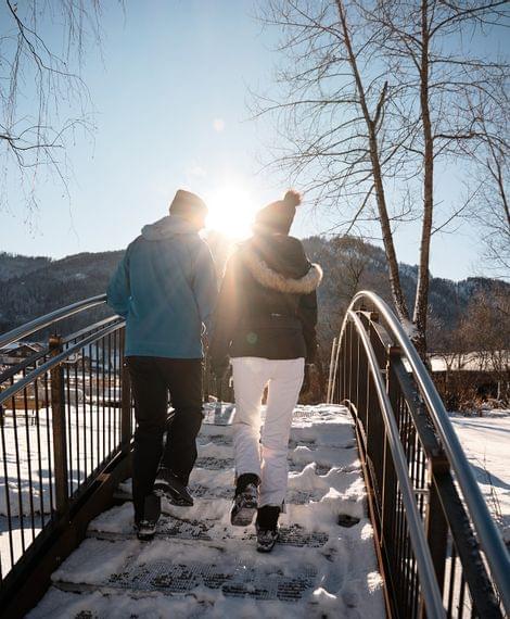 Paar geht über verschneite Brücke bei Sonnenschein in winterlicher Berglandschaft
