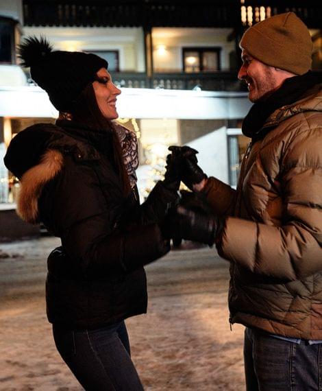 Lächelndes Paar in Winterkleidung hält Hände vor beleuchtetem Hotel im Schnee
