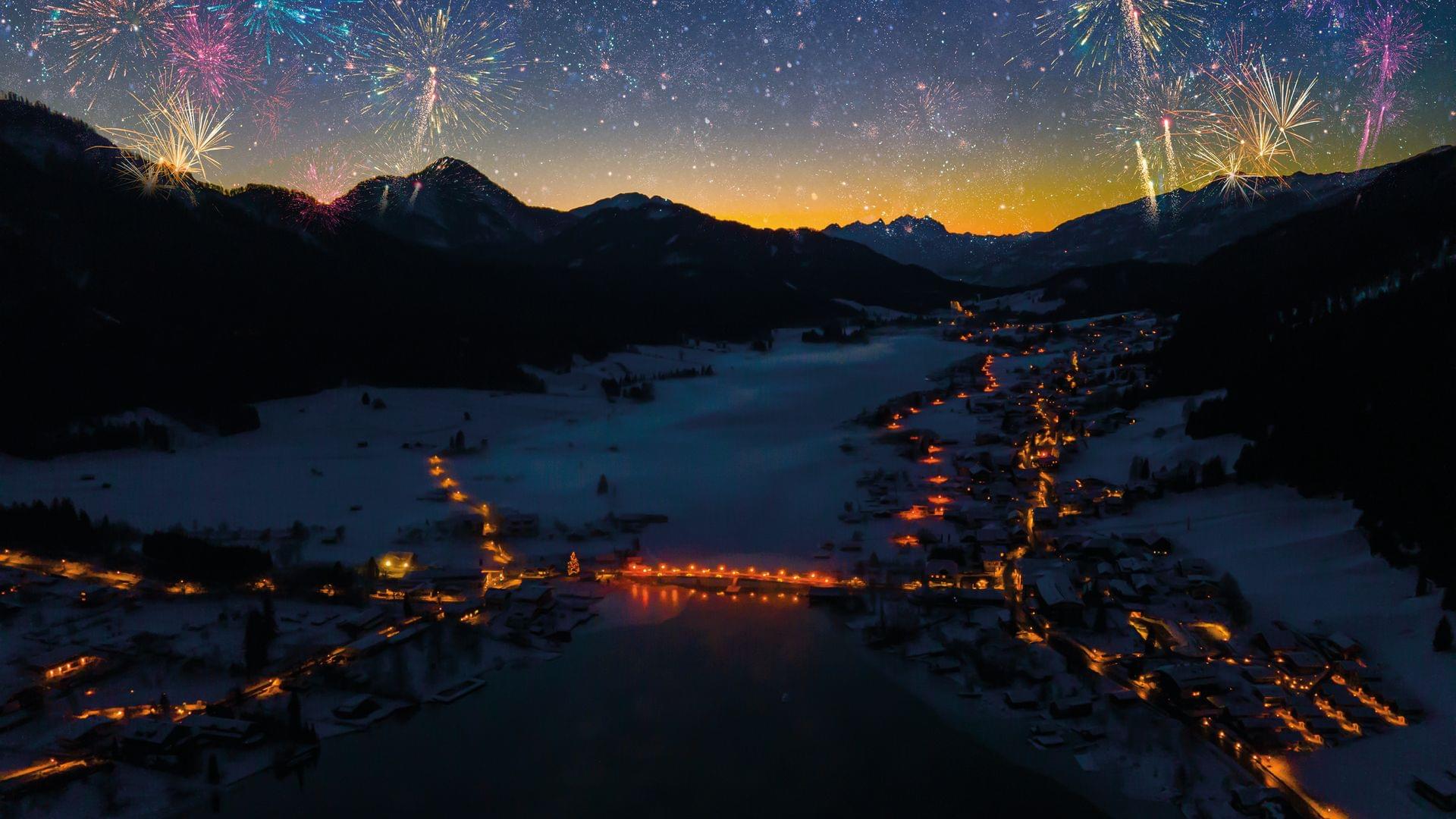 Feuerwerk über verschneitem Alpenort mit Lichtern entlang eines zugefrorenen Sees bei Nacht