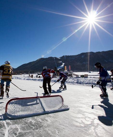 Eishockeyspiel im Freien bei Sonnenschein mit verschneiten Bergen im Hintergrund
