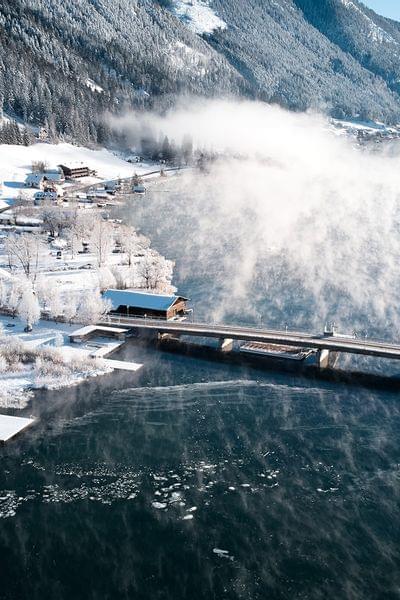 Winterliches Alpenpanorama mit verschneitem Dorf am See und dampfender Wasseroberfläche