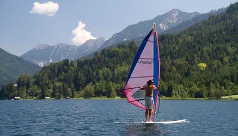 Mann beim Windsurfen auf einem See vor Alpenkulisse und bewaldeten Hügeln