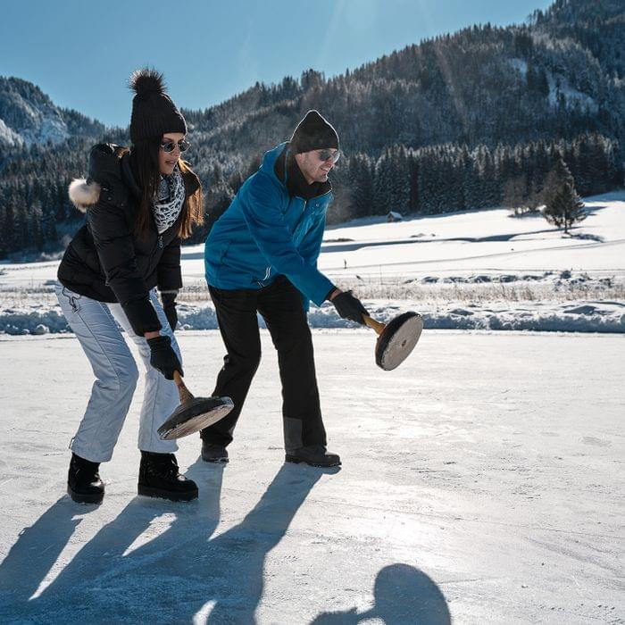 Zwei Personen spielen Eisstockschießen auf gefrorenem See in winterlicher Berglandschaft