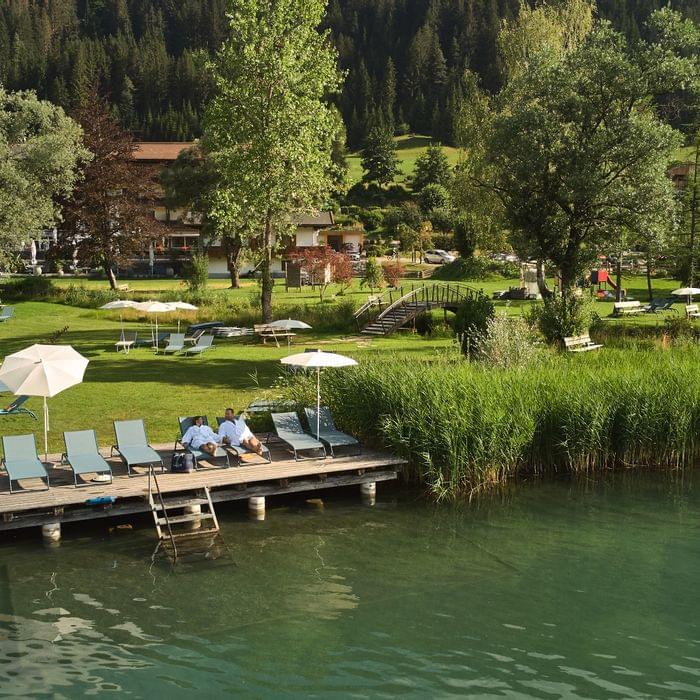 Sonnenliegen und Sonnenschirme am Badesteg eines Sees mit grüner Wiese und Bergkulisse