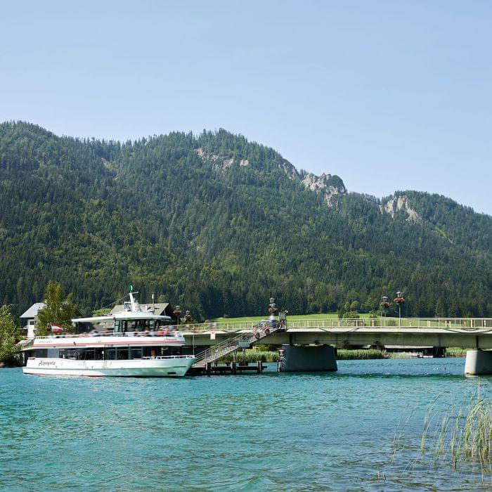 Ausflugsschiff auf türkisblauem See vor Brücke und bewaldeten Bergen im Sommer