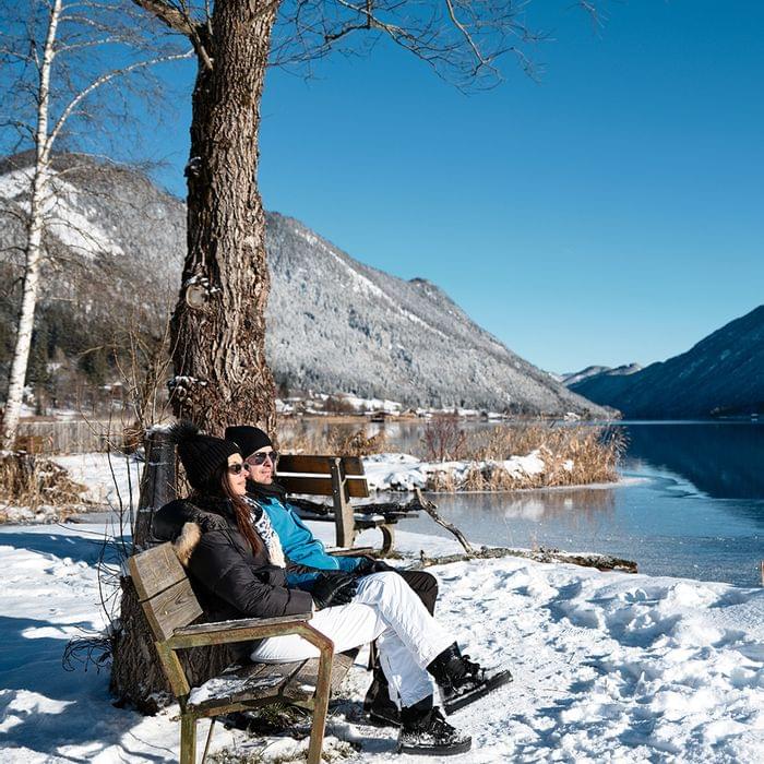 Paar sitzt im Winter auf einer Bank am verschneiten See in den Alpen bei klarem Himmel