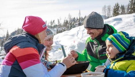 Familie isst gemeinsam im Schnee mit Winterkleidung bei sonnigem Wetter