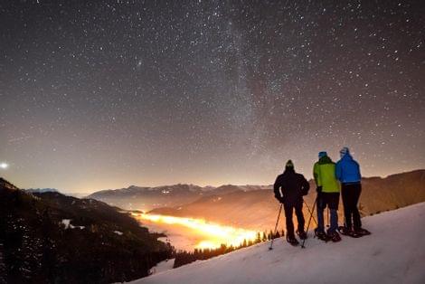 Drei Schneeschuhwanderer blicken nachts auf ein beleuchtetes Tal unter sternenklarem Himmel in den Alpen