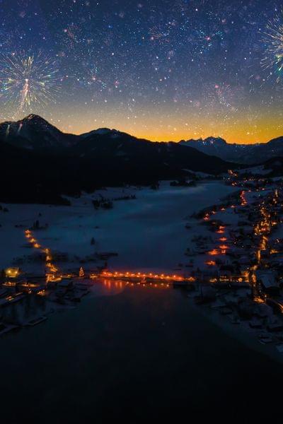 Feuerwerk über verschneitem Alpenort mit Lichtern entlang eines zugefrorenen Sees bei Nacht