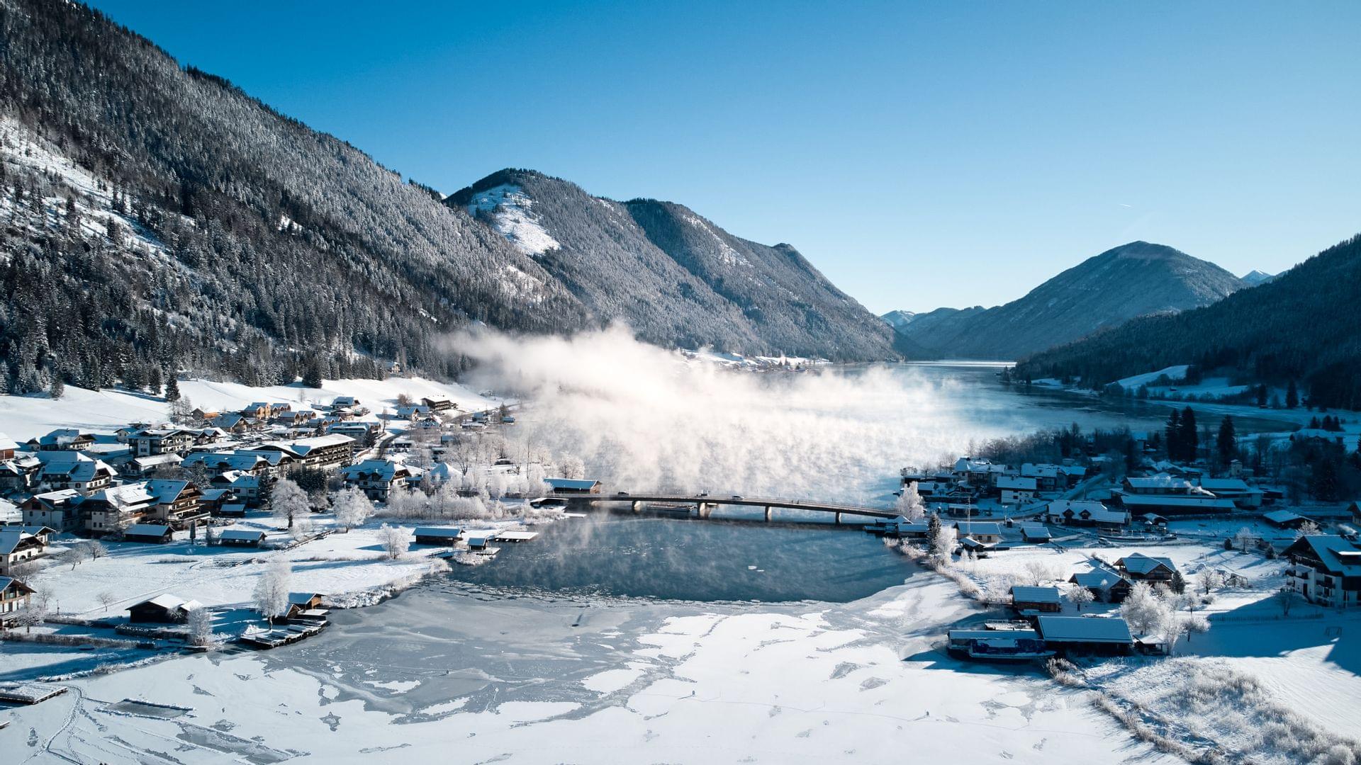Winterlandschaft am zugefrorenen See mit Brücke und schneebedecktem Alpendorf in den Bergen