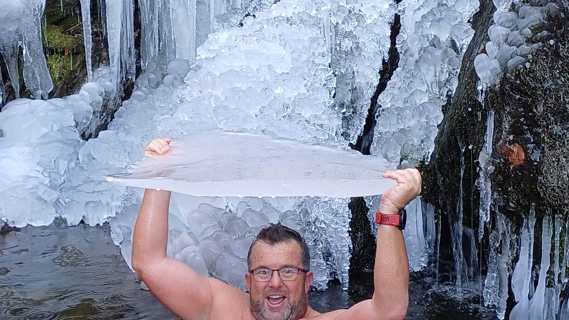 Mann sitzt in eiskaltem Wasser vor gefrorenem Wasserfall und hält eine große Eisscholle über dem Kopf