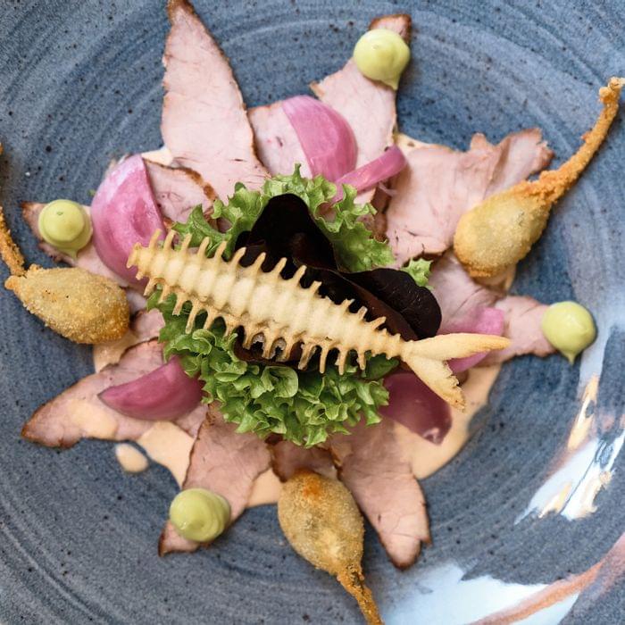 Kreativ angerichtetes Carpaccio mit frittiertem Gemüse, Salat und cremigen Tupfen auf blauem Teller
