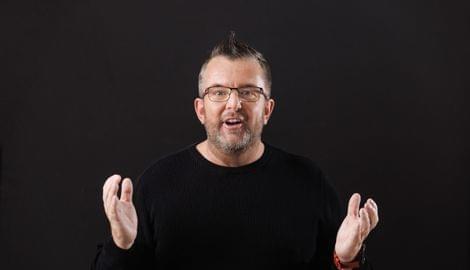 Mann mit Brille und Bart spricht gestikulierend vor schwarzem Hintergrund