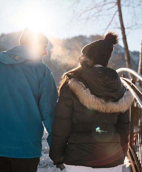 Paar in Winterkleidung geht über eine verschneite Brücke bei Sonnenschein in den Bergen
