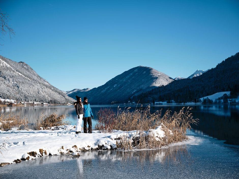 Winterwanderer am verschneiten See mit Bergen im Hintergrund bei klarem Himmel