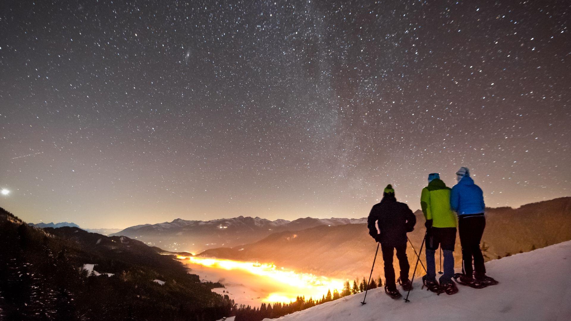 Drei Schneeschuhwanderer blicken bei Nacht auf ein beleuchtetes Tal unter einem klaren Sternenhimmel