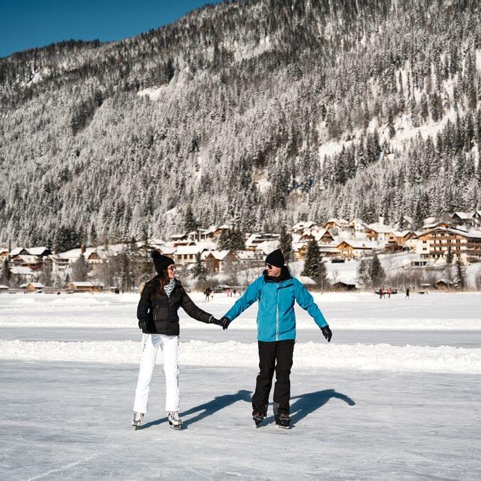 Paar beim Eislaufen auf zugefrorenem See vor verschneitem Bergdorf in den Alpen