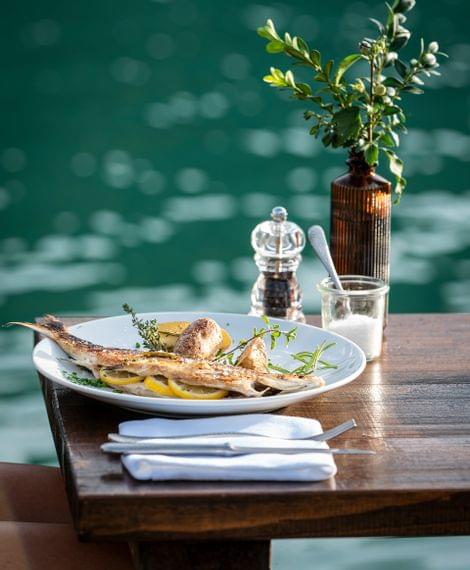 Gegrillter Fisch mit Kartoffeln und Zitrone auf einem Holztisch am Wasser serviert