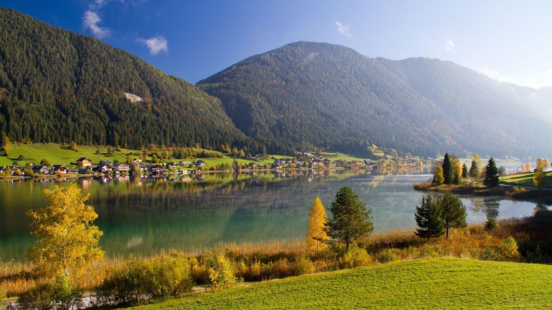 Alpensee mit klarer Spiegelung, umgeben von grünen Hügeln und herbstlichen Bäumen