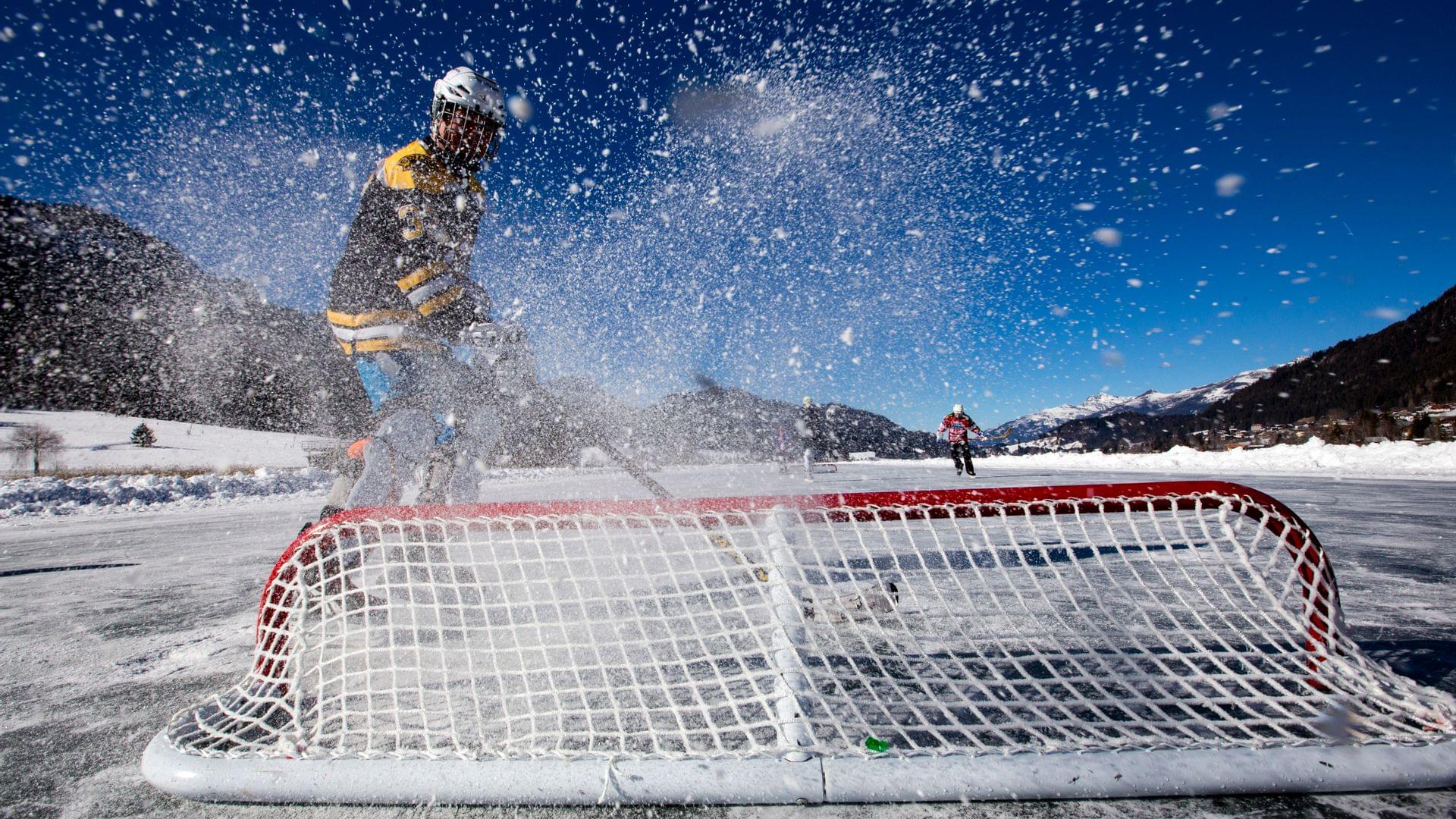 Eishockeyspieler schießt auf ein Tor auf einem zugefrorenen See in winterlicher Berglandschaft