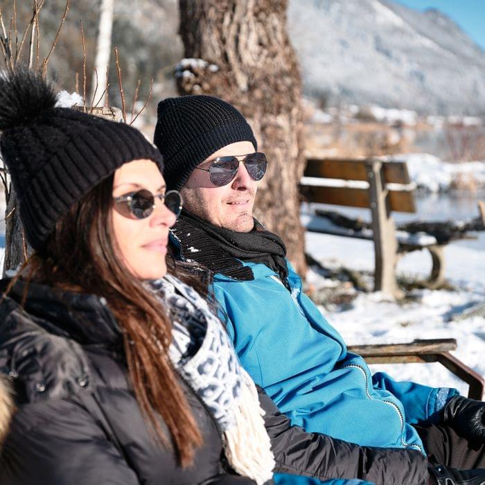 Paar in Winterkleidung entspannt auf Bank in verschneiter Alpenlandschaft in der Sonne