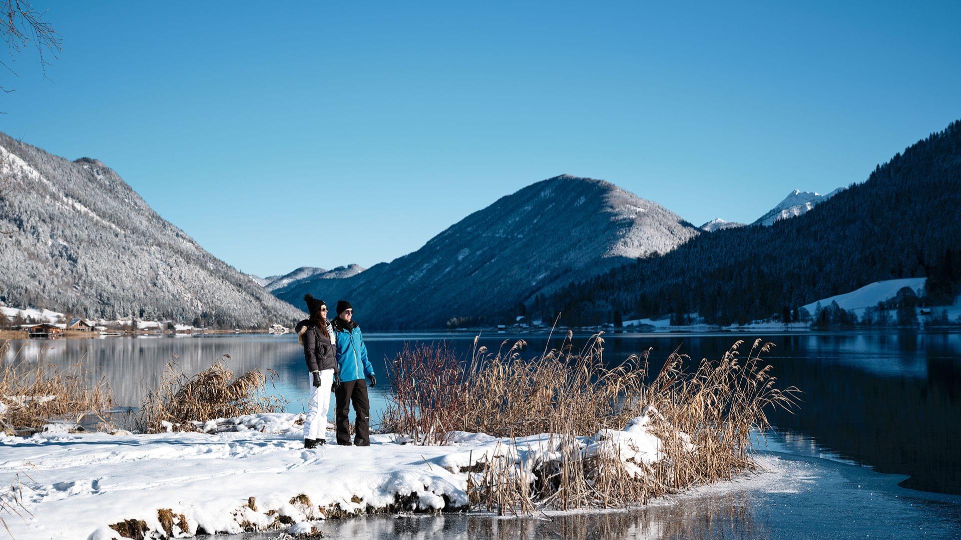 Winterwanderer am verschneiten See mit Bergen im Hintergrund bei klarem Himmel