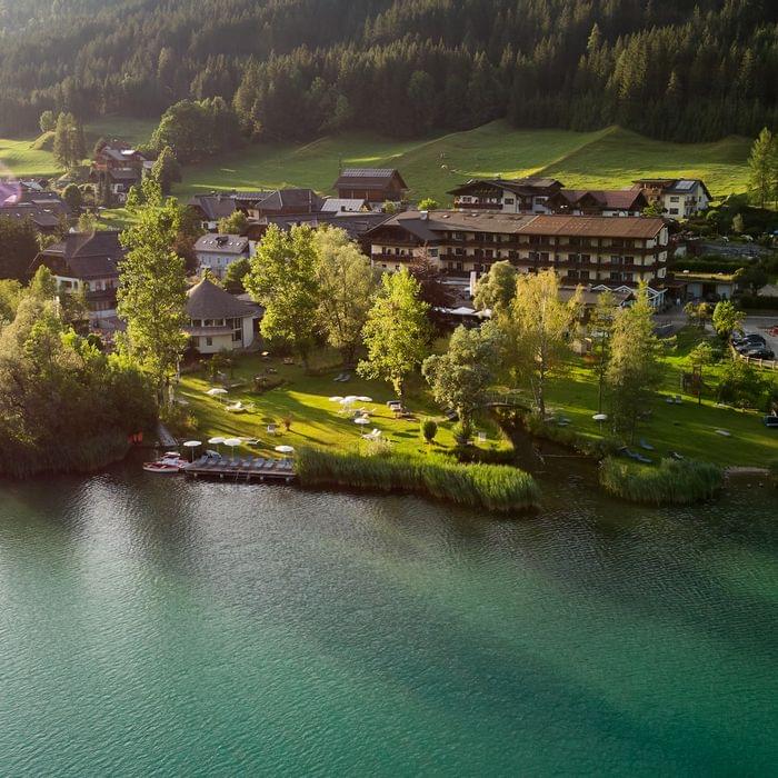 Luftaufnahme eines Hotels am Seeufer mit Liegewiese und umliegenden Bergen im Sonnenlicht