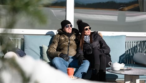 Paar in Winterkleidung entspannt auf Sofa in der Sonne mit Tee und Schnee im Vordergrund
