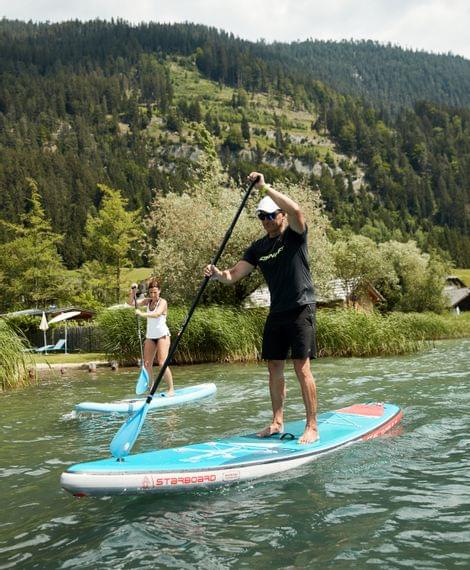 Zwei Personen beim Stand-Up-Paddling auf einem klaren See vor bewaldeter Bergkulisse