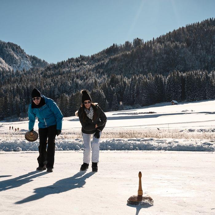 Zwei Personen spielen Eisstockschießen im verschneiten Alpenpanorama bei Sonnenschein