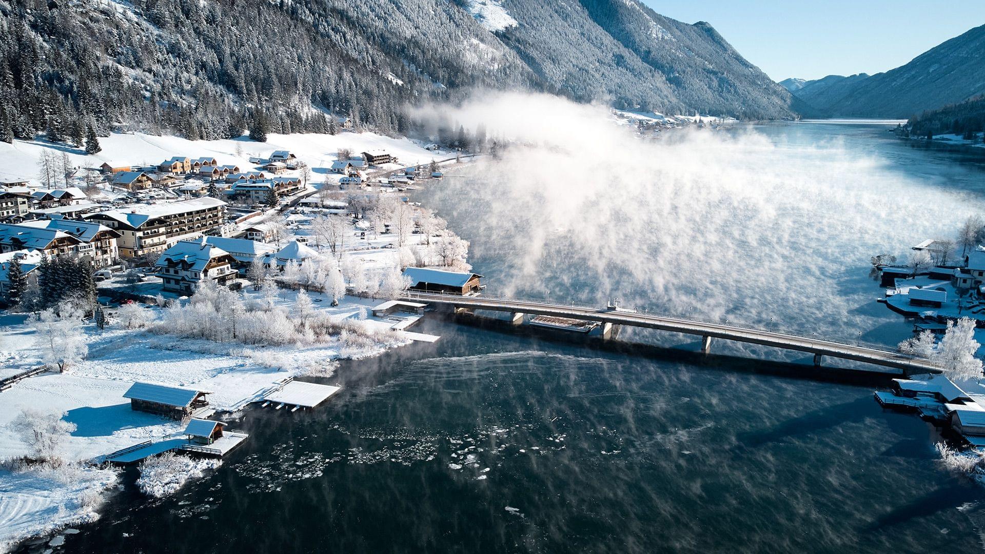 Winterliches Alpenpanorama mit verschneitem Dorf am See und dampfender Wasseroberfläche