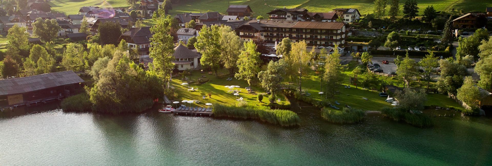 Luftaufnahme eines Hotels am Seeufer mit Liegewiese und umliegenden Bergen im Sonnenlicht