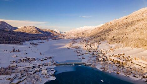 Winterlandschaft mit verschneiten Bergen, gefrorenem See und Dorf in den Alpen bei Sonnenaufgang