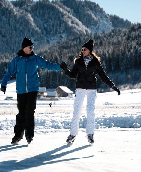 Paar beim Eislaufen im Freien vor schneebedeckten Bergen und winterlichem Wald