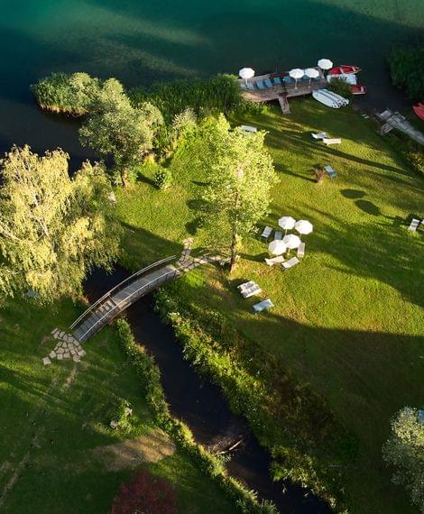 Grüne Liegewiese mit Sonnenschirmen und Steg an einem klaren Badesee aus der Vogelperspektive